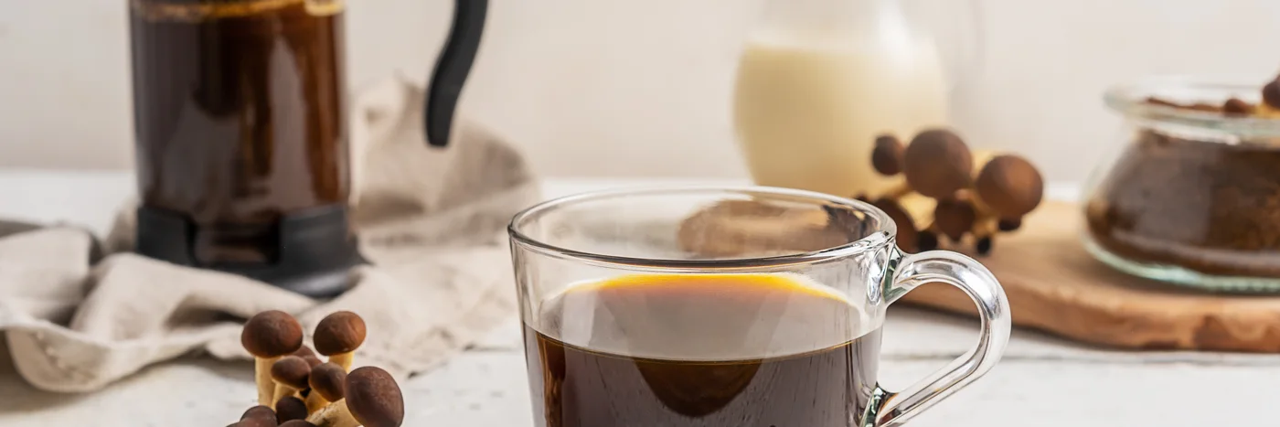 A glass cup of black mushroom coffee beside a stovetop pot and a scattering of dried mushrooms on a pale wooden surface.
