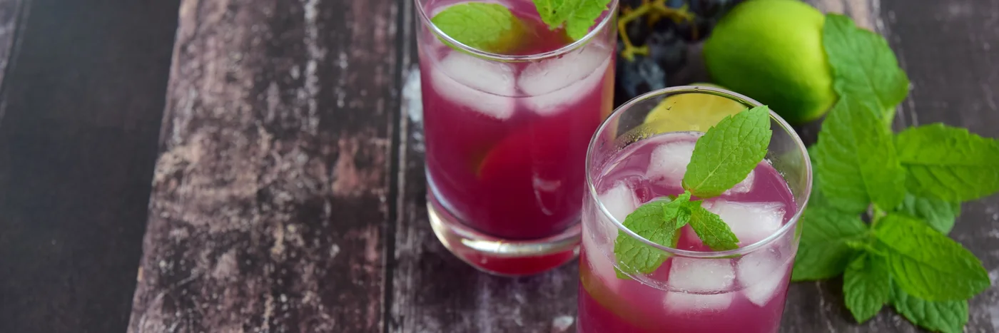 Two glasses of deep berry-coloured botanical soda over ice with fresh mint, on dark weathered wood.
