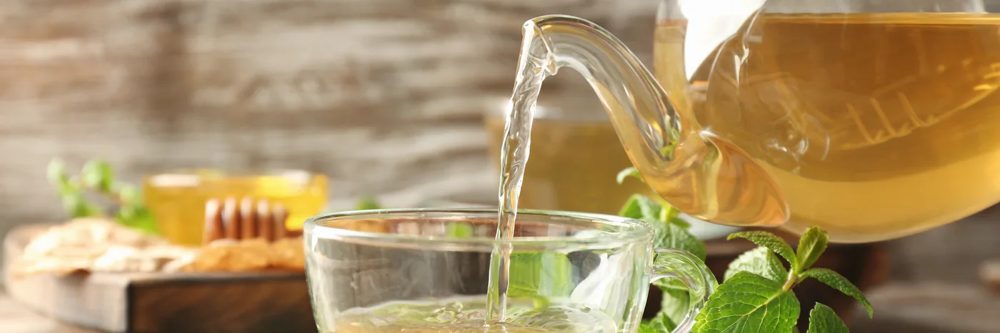Lemon balm tea being poured from a glass pot into a cup, with fresh lemon balm leaves alongside.
